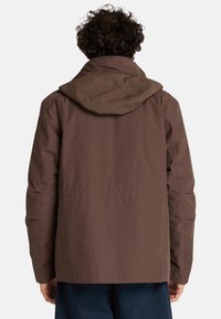 Giacca impermeabile marrone con cappuccio, caratterizzata da un design semplice e dritto e una texture liscia. Sono visibili i polsini regolabili.