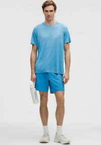 Niebieska koszulka sportowa z teksturowanym wzorem, w połączeniu z jednolitymi niebieskimi szortami. Model trzyma biały bidon. Całość dopełniają sneakersy i skarpety typu crew.