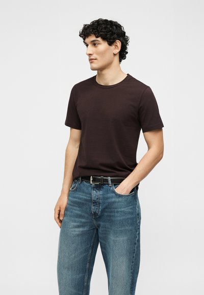 Άνδρας με σγουρά μαλλιά, που φοράει ένα σκούρο καφέ κοντομάνικο μπλουζάκι, μπλε τζιν και μαύρη ζώνη, στέκεται με το ένα χέρι στην τσέπη.