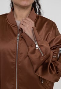 Bruine satijnen bomberjack met een geribde kraag, met meerdere ritsen op de mouwen en een gestroomlijnd ontwerp. Zachte textuur en slim fit stijl.
