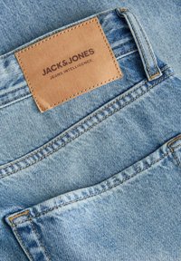 Lysblå denimjeans med en tan læderpatch med præget branding på bagsiden, der fremviser synlige syninger og lomme detaljer.