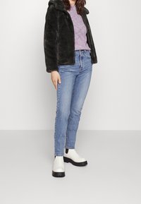 Veste en fausse fourrure vert foncé, pull violet clair, jean bleu et bottines blanches à semelles noires. Tenue décontractée avec des couches texturées.