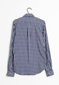 Camicia a maniche lunghe a quadri blu navy e bianca, con colletto, bottoni ai polsini e orlo leggermente curvato, appesa a un gancio nero.