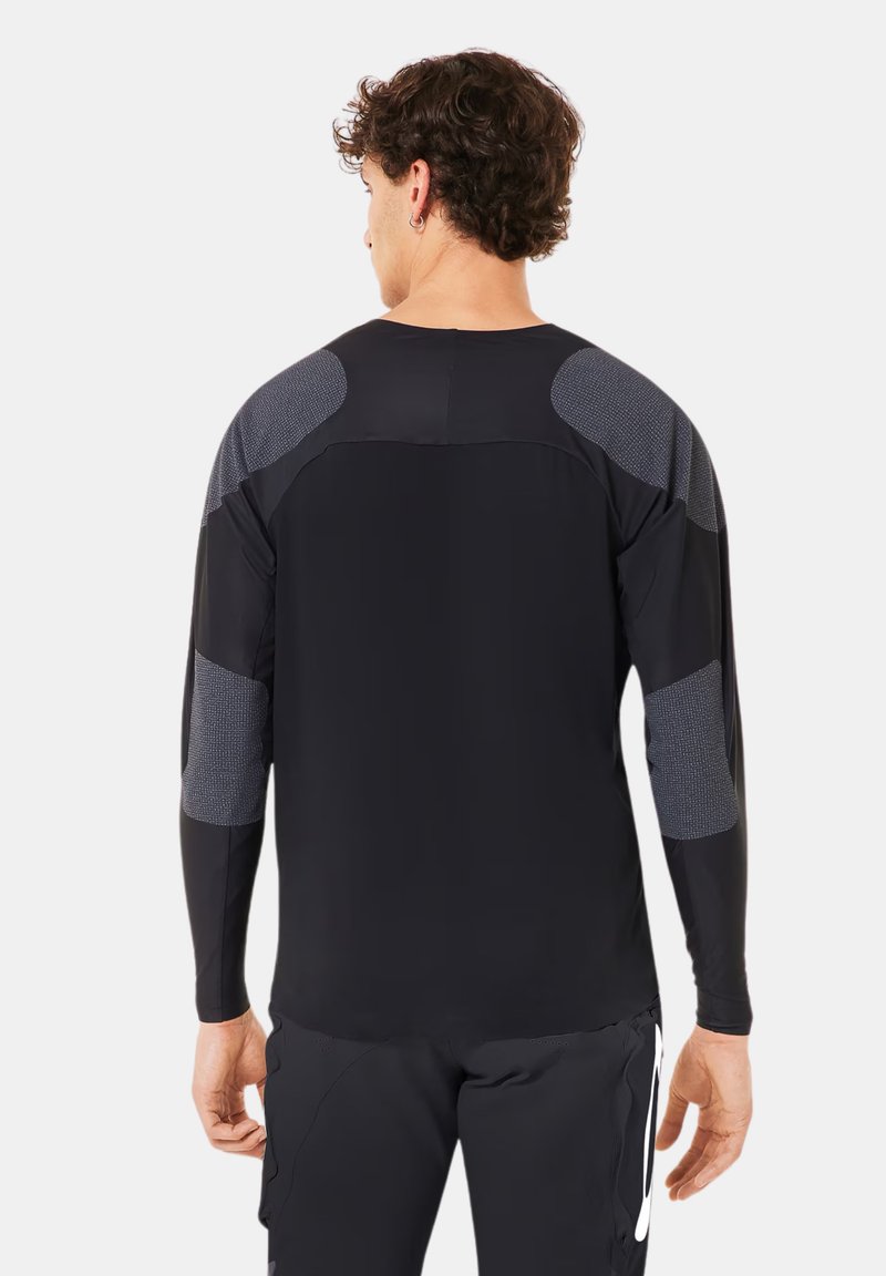 Homme portant un maillot de sport noir à manches longues avec des patchs gris texturés aux épaules et aux coudes, et un pantalon noir avec un logo blanc, vu de dos.