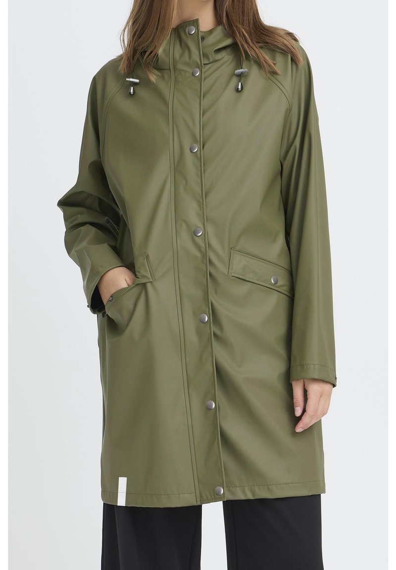 Femme portant un imperméable vert olive à boutons-pression et capuche avec cordon, d'une longueur jusqu'aux genoux, une main dans la poche, debout devant un fond blanc.