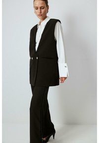 Black sleeveless vest with a V-neck and decorative buttons, paired with matching trousers featuring side button accents. White shirt underneath.