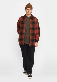 Rode en zwarte geruite flanellen jas met twee borstzakken, ritsluiting, gedragen over een bruine shirt; gecombineerd met zwarte broek en sneakers.