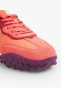 Zapatilla deportiva con parte superior de ante y sintético en tonos de rosa y naranja, con una suela exterior de goma texturizada morada con estudios hexagonales.