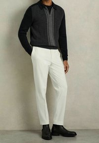 Černý a šedý pletený svetr na zip s texturovaným vzorem, doplněný bílými kalhotami na míru a černými kotníkovými botami.