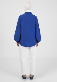 Camisa azul de manga larga con cuello, puños fruncidos y dobladillo curvado. Combinada con pantalones blancos y zapatillas azules. Textura de tela suave.