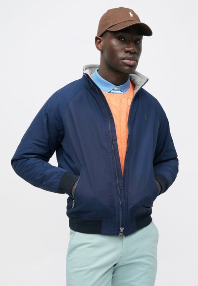 Navy bomber jacket with gray lining and ribbed cuffs, worn over an orange knit sweater, paired with light blue collared shirt and mint pants.