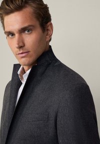 Giacca in misto lana grigio scuro con colletto alto, taschino sul petto, e una texture sottile, sovrapposta a una camicia bianca con motivo a quadretti.