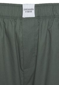 Vihreät kangashousut, joissa on joustava vyötärö, sivutasku ja tikattu yksityiskohta. Tarrassa lukee "COPENHAGEN STUDIOS". Sujuva pinta.