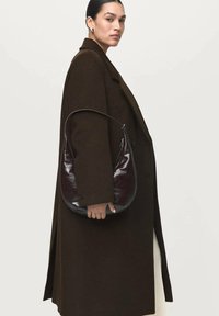 Abrigo de lana marrón sobre un conjunto crema, sosteniendo un bolso liso de color burdeos oscuro con forma curva y una correa corta.