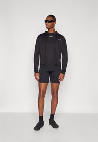 Schwarze Sportjacke mit Reißverschluss, figurbetonter Unterzieher und Shorts. Glatte Textur und atmungsaktives Material. Begleitet von dunklen Sonnenbrillen und Schuhen.