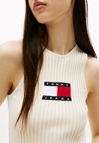 Ærmeløs ribbet top i beige med rund halsudskæring, der har et broderet Tommy Jeans-logo i rød, hvid og sort på forsiden.