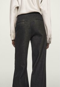 Mørke, vide bukser lavet af denim. Har to baglommer og en subtil glans, parret med en lysfarvet langærmet bluse.