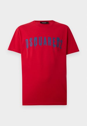 Jeune homme portant un T-shirt rouge Dsquared2, un pantalon noir ample et des chaussures rouges, debout devant un fond blanc uni.