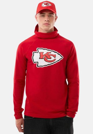 Młody mężczyzna ubrany w czerwoną bluzę Kansas City Chiefs i pasującą czerwoną czapkę, stojący z jedną ręką w kieszeni na jednolitym tle.