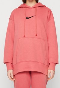 Person som bär en överdimensionerad korallrosa Nike-hoodie och matchande sweatpants, står med avslappnade händer vid sidorna.