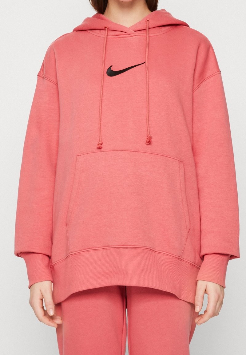 Person som bär en överdimensionerad korallrosa Nike-hoodie och matchande sweatpants, står med avslappnade händer vid sidorna.