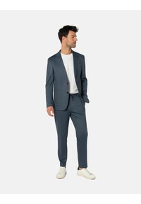 Maßgeschneiderter blauer Anzug bestehend aus einem Zweiknopf-Sakko und schmal zulaufenden Hosen mit dezentem Strukturmuster. Dazu kombiniert mit einem weißen T-Shirt und hellen Sneakers.