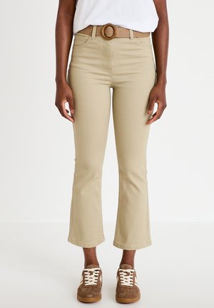 Person wearing beige cropped flare pants with a woven belt and brown suede sneakers with white laces against a plain background.