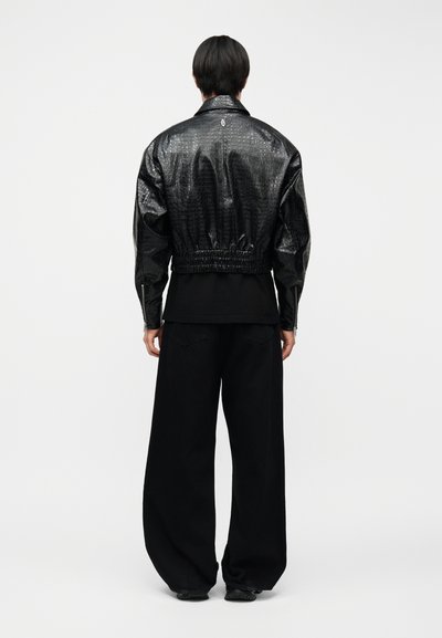 Άτομο που στέκεται με την πλάτη εμφανή, φοράει cropped μαύρο δερμάτινο σακάκι με υφή και φαρδιά μαύρη παντελόνια.