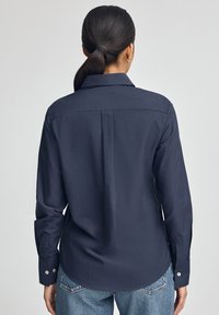 Granatowa, długa koszula z guzikami i kołnierzykiem, z szwami na plecach i detalami na mankietach. W połączeniu z jasnymi dżinsami.