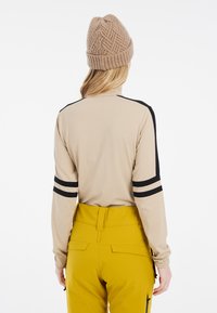 Beige Langarmoberteil mit schwarzen Streifen an den Ärmeln, kombiniert mit hochgeschnittenen gelben Hosen. Das Outfit wird von einem strukturierten braunen Strickmütze vervollständigt.
