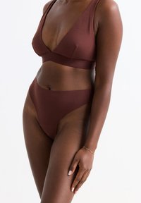 Vrouw in een bijpassende set van een donkerbruine bralette en hooggeknoopte onderbroek met een gouden armband om haar linkerpols, tegen een witte achtergrond.