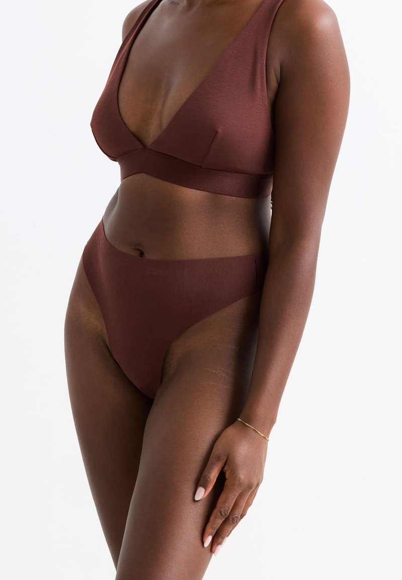 Vrouw in een bijpassende set van een donkerbruine bralette en hooggeknoopte onderbroek met een gouden armband om haar linkerpols, tegen een witte achtergrond.