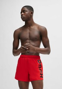 Red swim shorts with an elastic waistband, featuring a black "HUGO" logo printed on the left side. Smooth fabric with a classic cut.