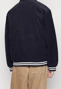 Uomo che indossa una giacca di velluto a corda blu navy con polsini e orlo a righe, abbinata a pantaloni di velluto a corda marrone chiaro, visto da dietro.