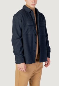 Veste chemise en mélange de laine navy avec deux poches poitrine, fermeture à boutons et col pointu. Portée sur un col roulé beige. Texture lisse.