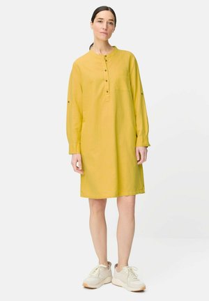 Kvinde stående iført en gul langærmet knapbåret kjole og hvide sneakers mod en ensartet lys baggrund.