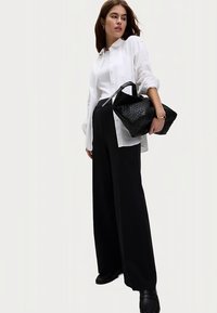 Sort flettet håndtaske, hvid skjorte og sorte bukser med vide ben. Outfit'et har et struktureret design og glatte teksturer. Fodtøjet er sort og tekstureret.