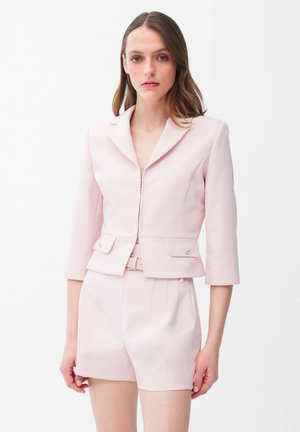 Kvinde iført en sart lyserød skræddersyet blazer med trekvartlange ærmer og matchende højtaljede shorts mod en hvid baggrund.