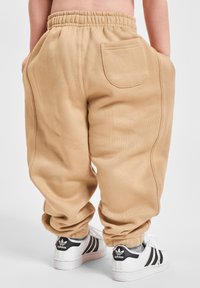 Kind trägt beige Trainingshosen mit elastischen Bündchen und einer Gesäßtasche, kombiniert mit weißen Adidas Sneakers mit schwarzen Streifen.