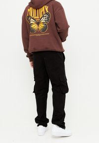 Felpa con cappuccio marrone con una grande grafica di una farfalla e testo sulla schiena, abbinata a pantaloni cargo neri e sneakers bianche.