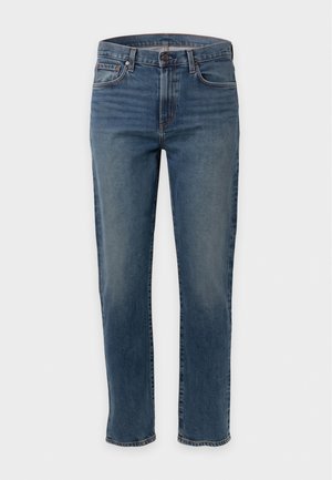 Homme debout portant une chemise boutonnée vert olive, un t-shirt beige, un jean bleu et des chaussures en cuir noires sur un fond blanc uni.