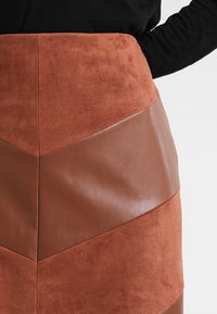 Gros plan sur une jupe en suède brun et en cuir avec un motif en chevrons, portée avec un haut à manches longues noir.