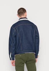 Chaqueta de mezclilla de color azul oscuro, con costuras en contraste y un cuello de borreguillo blanco. Cuenta con bolsillos laterales y detalles de botones en los puños.