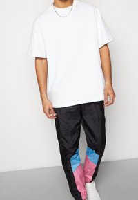 Vit oversized t-shirt med svarta träningsbyxor som har rosa och blå geometriska detaljer. Modellen står mot en neutral bakgrund.