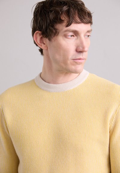 Gelbes geripptes Strickpullover mit einem kontrastierenden beigen Rundhalsausschnitt. Fein strukturiert mit vertikalen Streifen, die ein tailliertes Design betonen.