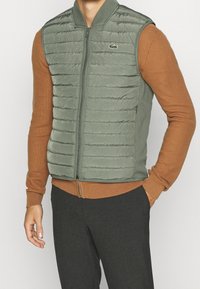 Green lightweight puffer vest with horizontal quilted pattern, zip front, and stand collar. Worn over a brown long-sleeve shirt.