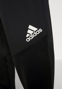 Pantalones deportivos negros con una textura suave, con un logotipo blanco de Adidas en el lado izquierdo cerca de la cadera, diseñados para comodidad y flexibilidad.