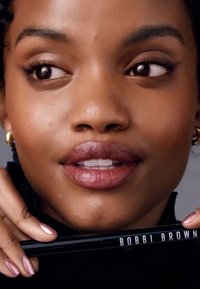 Svart eyeliner blyant med teksten "BOBBI BROWN", holdt i en hånd. Subjektet har en jevn hudfarge, fyldige lepper med glans, og har på seg en mørk topp.