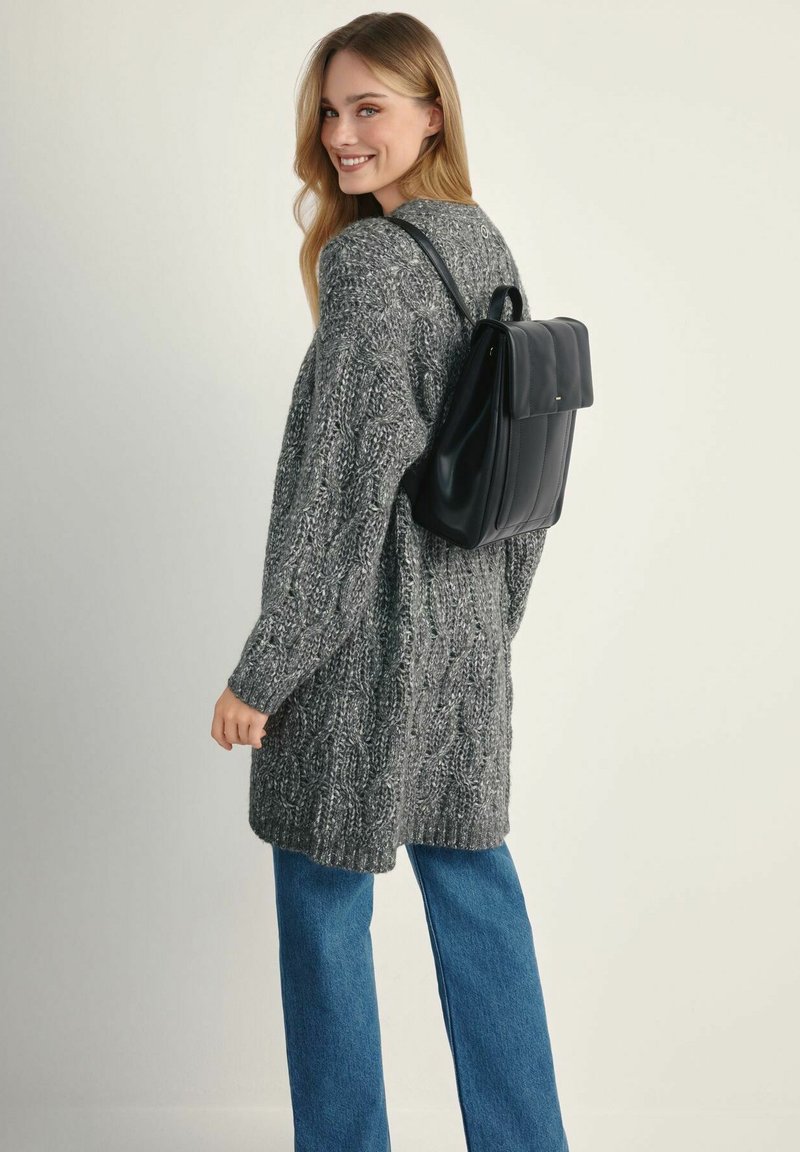 Czarny skórzany plecak o zorganizowanej konstrukcji, noszony na plecach. Modelka w szarym, dzianinowym kardiganie i niebieskich szerokich spodniach.