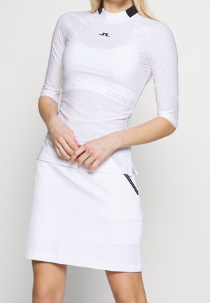 Nainen, joka pukeutunut istuvaan valkoiseen kolmivartiseen puseroon ja valkoiseen hameeseen, jossa on musta logo rinnassa ja musta somiste hameen taskussa.